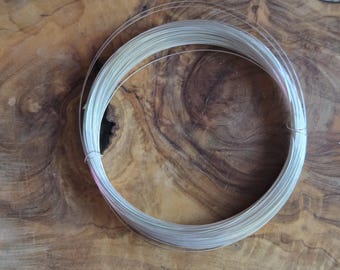 May include: A coiled bundle of thin, silver-colored wire. The wire is tightly wound into a circular shape, with the individual strands visible. The background is a wooden surface with a natural grain pattern. The wire appears to be for jewelry making or crafts.
