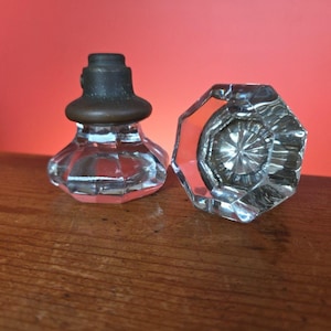 May include: Two antique glass door knobs. One knob is upright with a dark metal top and a clear, faceted glass base. The other knob is lying on its side, showcasing its multifaceted design. The background is a solid red.