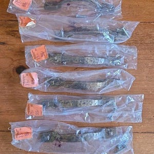 Może przedstawiać: Sześć antycznych, brązowych uchwytów do szafek, każdy zapakowany osobno w przezroczysty plastik. Uchwyty mają młotkowaną fakturę i są zaprojektowane z zakrzywionym uchwytem. Tło to drewniana powierzchnia.