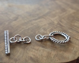 May include: A silver toggle clasp, featuring a bar with a beaded edge and an oval ring with a twisted rope design. The clasp is open, showing the two separate pieces against a wooden surface.