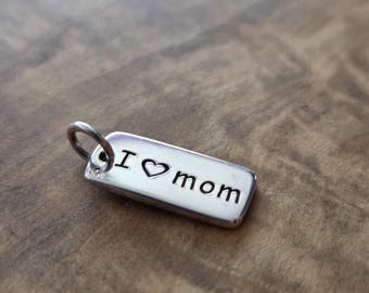 May include: A rectangular silver pendant with the words "I ♥ mom" engraved on it. The pendant has a small loop at the top for attaching to a chain or bracelet. The background is a wooden surface.
