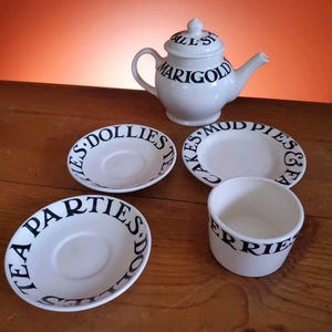 May include: A white teapot with black lettering that reads "All's Fair in Love and Marigold" and four white saucers with black lettering. The saucers read "Tea Parties Doll's Tea", "Cakes 'n' Mud Pies & Tea", "Tea Parties Doll's Tea", and "Berries & Tea".