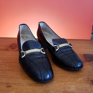 May include: A pair of black leather loafers with gold-colored accents. The shoes have a rounded toe and a small heel. The gold accent is a decorative bar across the top of the shoe. The shoes are on a wooden surface.