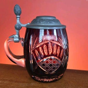 May include: A ruby red glass beer stein with a decorative cut glass design. The stein has a metal hinged lid and handle. The handle is clear glass. The stein is sitting on a wooden surface.