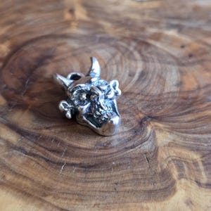 May include: A small, silver-colored pendant in the shape of a horned skull. The skull has detailed features, including horns, eye sockets, and a prominent jawline. The pendant is designed with a loop for attaching to a chain or cord.