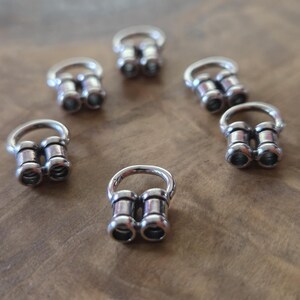 May include: Six silver-colored, double-flared ear gauges are arranged on a wooden surface. Each gauge has a cylindrical shape with a flared end and a ring for insertion. The gauges are designed for stretched ear piercings.