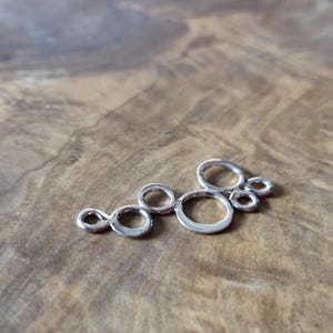 May include: A silver charm featuring a collection of linked, open circles. The charm is made of a shiny metal and is shown on a wooden surface. The design is abstract and could be used for jewellery making or other crafts.