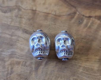 May include: Two silver-colored skull beads with detailed facial features, including eye sockets, a nose cavity, and a row of teeth. The beads are displayed on a wooden surface.
