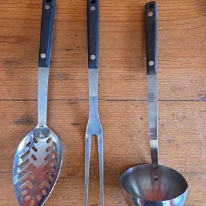 Könnte beinhalten: Drei Küchenutensilien aus Edelstahl mit schwarzen Griffen auf einer Holzoberfläche. Das Set besteht aus einem Schöpflöffel, einer Gabel mit zwei Zinken und einer Kelle. Die Griffe sind mit einer silbernen Niete befestigt.