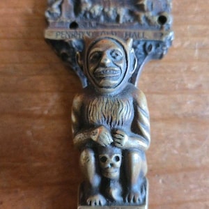May include: A bronze-colored metal sculpture of a seated imp holding a skull. The sculpture has the text "PENRHYN HALL" at the top and "PENRHYN IMP" at the bottom. The imp has a smiling face and pointed ears.
