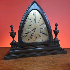 May include: A dark brown wooden clock with a triangular top and a gold-colored clock face. The clock features black Roman numerals and decorative finials on each side. The clock is set on a wooden surface.