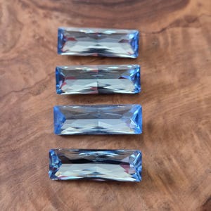 May include: Four rectangular, faceted, light blue gemstones are arranged vertically on a wooden surface. The gems have a clear, reflective quality, with sharp edges and a vibrant color. The wood grain provides a warm contrast to the cool tones of the gems.