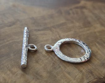 Può includere: Una chiusura a toggle color argento con finitura strutturata. La chiusura è composta da una barra e un anello, entrambi con piccoli anelli per il fissaggio. Il metallo ha un aspetto martellato, conferendogli un aspetto rustico. Ideale per la creazione di gioielli.