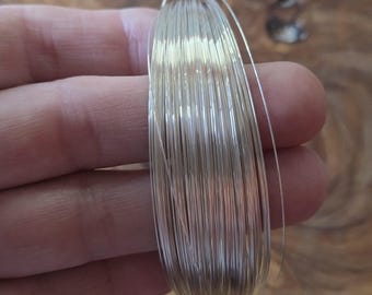 May include: A coil of thin, silver-colored wire, likely for jewelry making or crafts, is held in a hand. The wire is tightly wound, creating a compact bundle. The background is a blurred wooden surface.