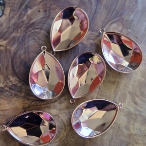 May include: Six teardrop-shaped pendants with faceted, reflective surfaces and a rose gold-colored metal frame. Each pendant has a small loop at the top for attaching to a chain or other jewelry component. The pendants are arranged on a wooden surface.