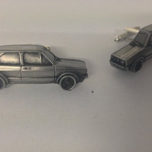 Puede incluir: Un par de gemelos de plata con forma de un coche clásico Volkswagen Golf. Los gemelos están detallados con un acabado plateado y ruedas negras.