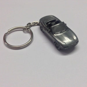 Puede incluir: Un llavero de metal plateado con un modelo detallado de un coche deportivo convertible. El coche tiene un interior negro y un acabado plateado.