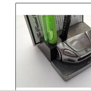 May include: A silver car-shaped pen holder with a green highlighter and a black pen. The holder is made of metal and has a brushed finish. The car is a Honda S2000.
