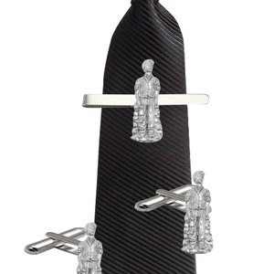 May include: A black tie with a silver tie bar and cufflinks featuring a silver figurine of a person. The figurine is wearing a long coat and holding a bag.