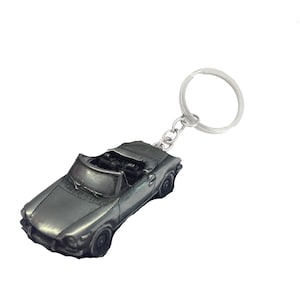 May include: A silver-colored metal keychain featuring a detailed model of a convertible car. The car has a black interior and a silver-colored body.