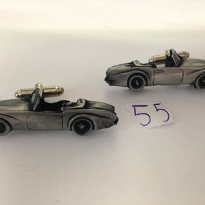 Puede incluir: Un par de gemelos de plata con forma de coches antiguos descapotables. Los gemelos están detallados con una parrilla y ruedas negras.