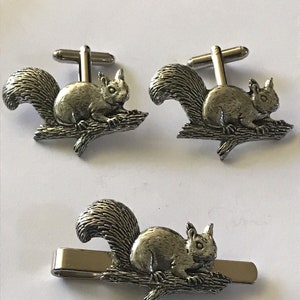 May include: A set of silver-toned cufflinks and a tie clip featuring a squirrel perched on a branch. The squirrel is detailed with a bushy tail and a small, pointed nose.