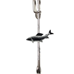 Puede incluir: Un broche de plata en forma de pez con cierre de seguridad. El pez está mirando hacia la derecha y tiene un diseño detallado.