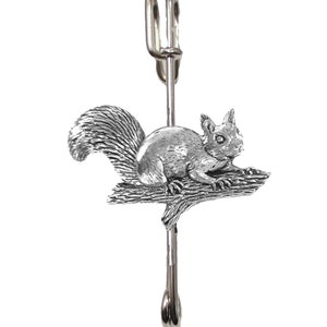 May include: A silver squirrel brooch with a detailed design of a squirrel sitting on a branch. The brooch has a safety pin closure.