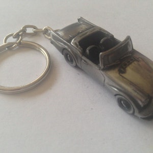 Puede incluir: Un llavero de metal plateado con un modelo detallado de un coche clásico descapotable. El coche tiene un interior negro y una carrocería plateada.
