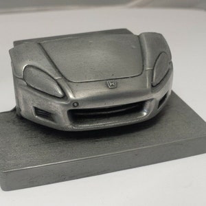 May include: A silver pewter model of a Honda S2000 sports car. The model is of the front of the car, showing the headlights, grille, and Honda logo.