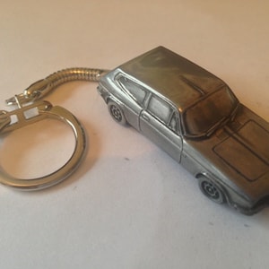 Puede incluir: Un llavero de metal plateado con un modelo detallado de un coche antiguo. El coche tiene una franja negra en el capó y un acabado plateado.