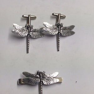 May include: A set of silver dragonfly cufflinks and a tie bar. The cufflinks and tie bar feature a detailed dragonfly design.