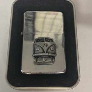 May include: A silver lighter with a raised, detailed design of a vintage Volkswagen van on the front. The lighter is in a black and silver tin.