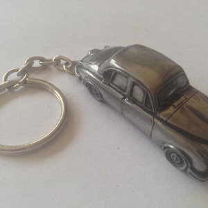 Può includere: Un portachiavi in metallo argentato con un modello dettagliato di un'auto d'epoca. L'auto ha una carrozzeria arrotondata e una griglia cromata.