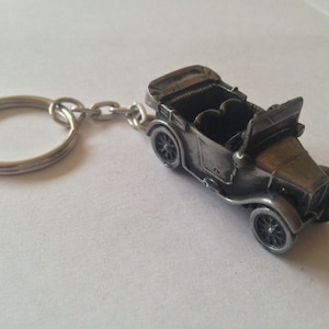 May include: A silver-coloured vintage car keyring. The miniature car has detailed features, including wheels, a convertible top, and a detailed interior. The keyring includes a metal ring and chain for attaching to keys or a bag.