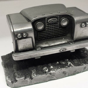 Puede incluir: Una escultura de metal plateado de la parte delantera de un Land Rover clásico. La escultura presenta faros detallados, una parrilla y un parachoques. Está montada sobre una base rectangular texturizada con un detalle de calavera, lo que sugiere un artículo de colección o decorativo.
