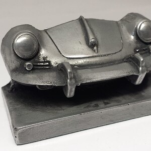 May include: A silver model of a classic Porsche 356, a vintage sports car, on a silver base. The model is detailed with headlights, a grille, and a curved hood.