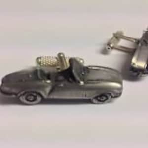 Könnte beinhalten: Ein Paar silberfarbene Manschettenknöpfe mit einem detaillierten Miniaturauto-Design. Das Auto ist ein Cabriolet mit schwarzem Verdeck und silbernen Rädern.