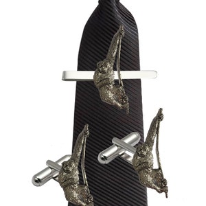 Puede incluir: Una corbata de rayas negras con un clip de corbata plateado que presenta un mono colgado de una vid. La imagen también muestra un par de gemelos plateados con el mismo diseño de mono.