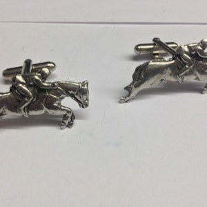 Puede incluir: Un par de gemelos de plata con un caballo y un jinete saltando un obstáculo. Los gemelos son detallados y realistas.