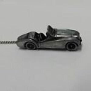 May include: A silver-colored, miniature replica of a classic convertible car with a detailed design. The car is attached to a silver-colored tie tack.