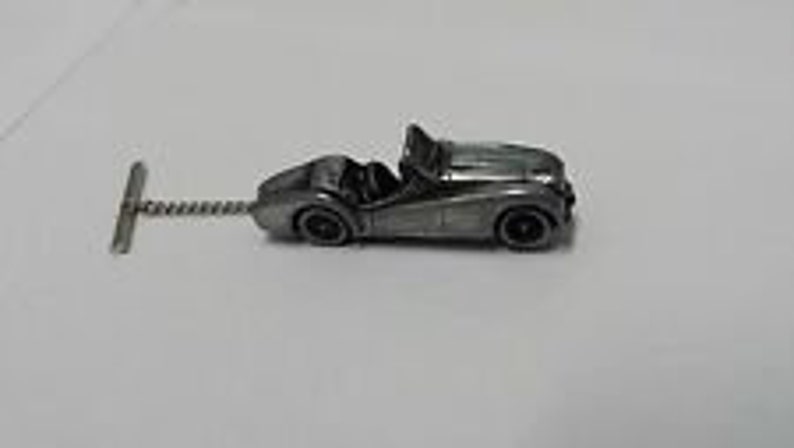 May include: A silver-colored, miniature replica of a classic convertible car with a detailed design. The car is attached to a silver-colored tie tack.