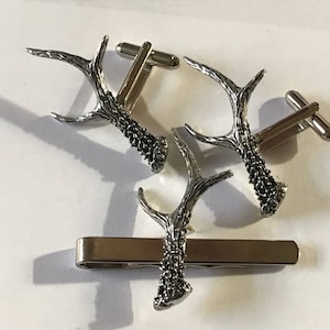 May include: A set of silver cufflinks and a tie clip featuring a detailed antler design. The cufflinks and tie clip are made of metal and have a polished finish.
