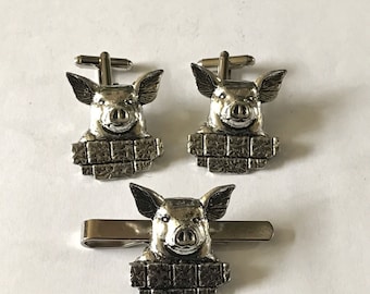 May include: A set of silver pig cufflinks and a tie clip. Each piece features a pig's head peeking over a brick wall.