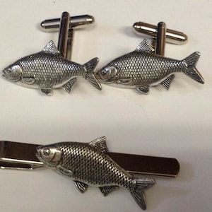 Könnte beinhalten: Ein Set silberner Fisch-Manschettenknöpfe und ein Krawattenclip. Die Fische sind detailliert und realistisch.