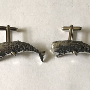 Puede incluir: Un par de gemelos de plata con un diseño detallado de cachalote. Las ballenas están mirando hacia la derecha y tienen una superficie texturizada.