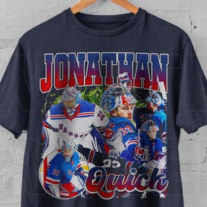 Può includere: Una t-shirt blu navy con una grafica in stile vintage del giocatore di hockey Jonathan Quick. La grafica mostra Quick con una divisa da hockey blu e rossa con il testo "Jonathan Quick" in un gradiente rosso e blu.