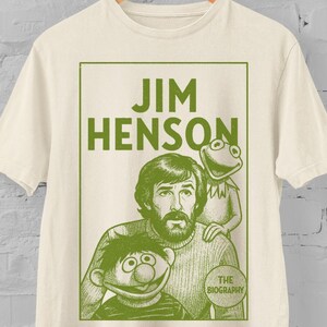 Puede incluir: Camiseta color crema con un gráfico verde de Jim Henson con la Rana Gustavo y Epi. El texto "JIM HENSON" está en la parte superior, con "THE BIOGRAPHY" en un círculo. La palabra "natural" está escrita en cursiva en la parte inferior.