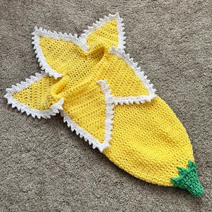 Può includere: Una copertina per neonati all'uncinetto a forma di limone. La copertina è gialla con un bordo bianco e una foglia verde in basso.