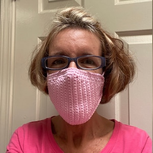 May include: A woman wearing a pink crocheted face mask and blue-striped glasses.